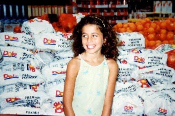 A young Charlotte smiling at a produce market, surrounded by fresh oranges and sacks of produce