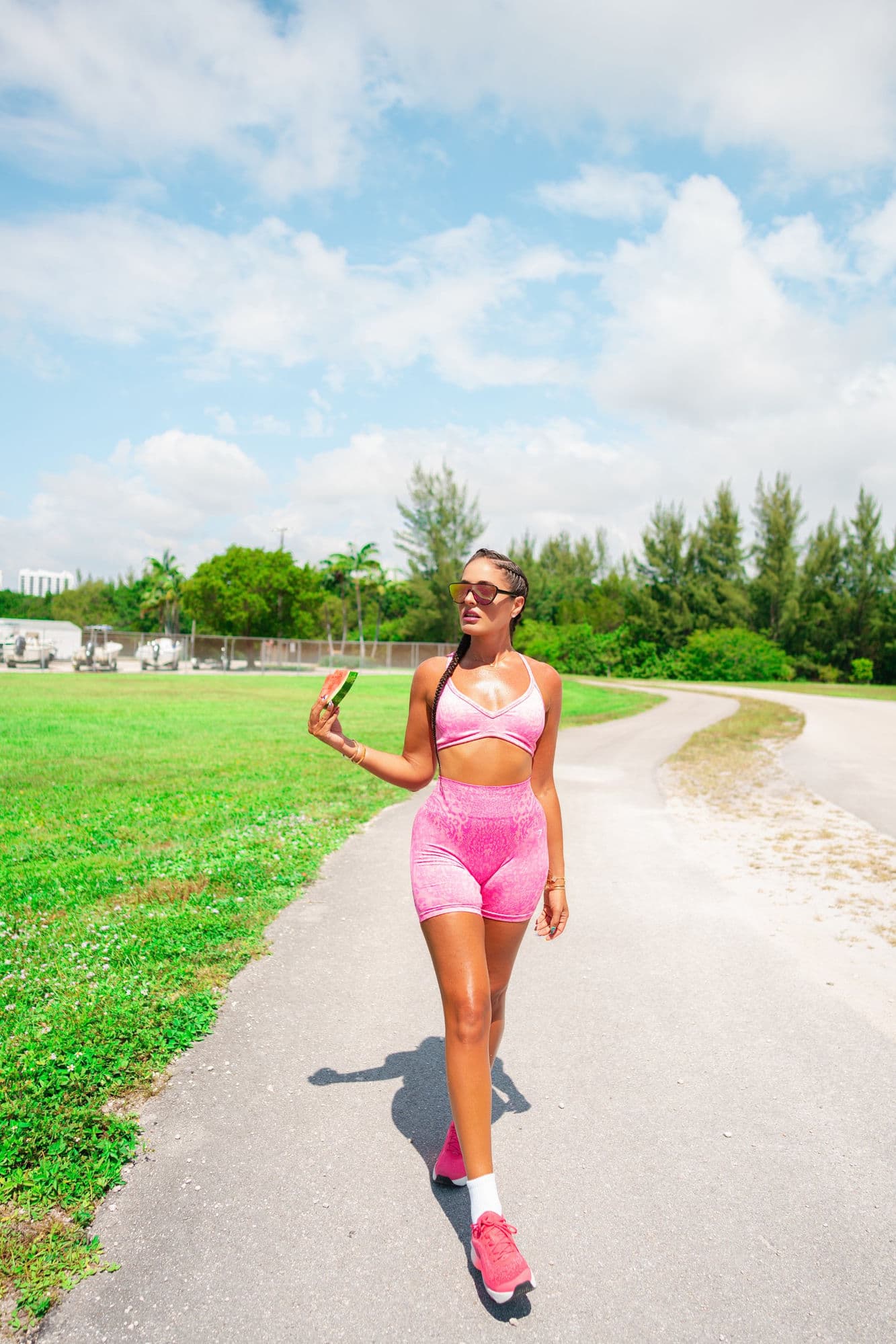 Charlotte walking a sunlit path in pink activewear, holding a slice of watermelon
