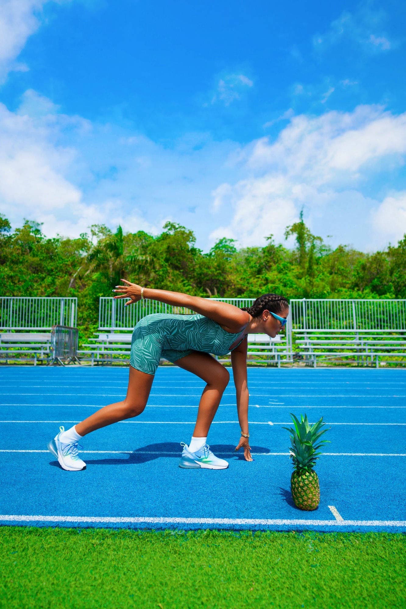 Charlotte in teal palm-print activewear in a sprint-start position on a blue running track, a pineapple at the line