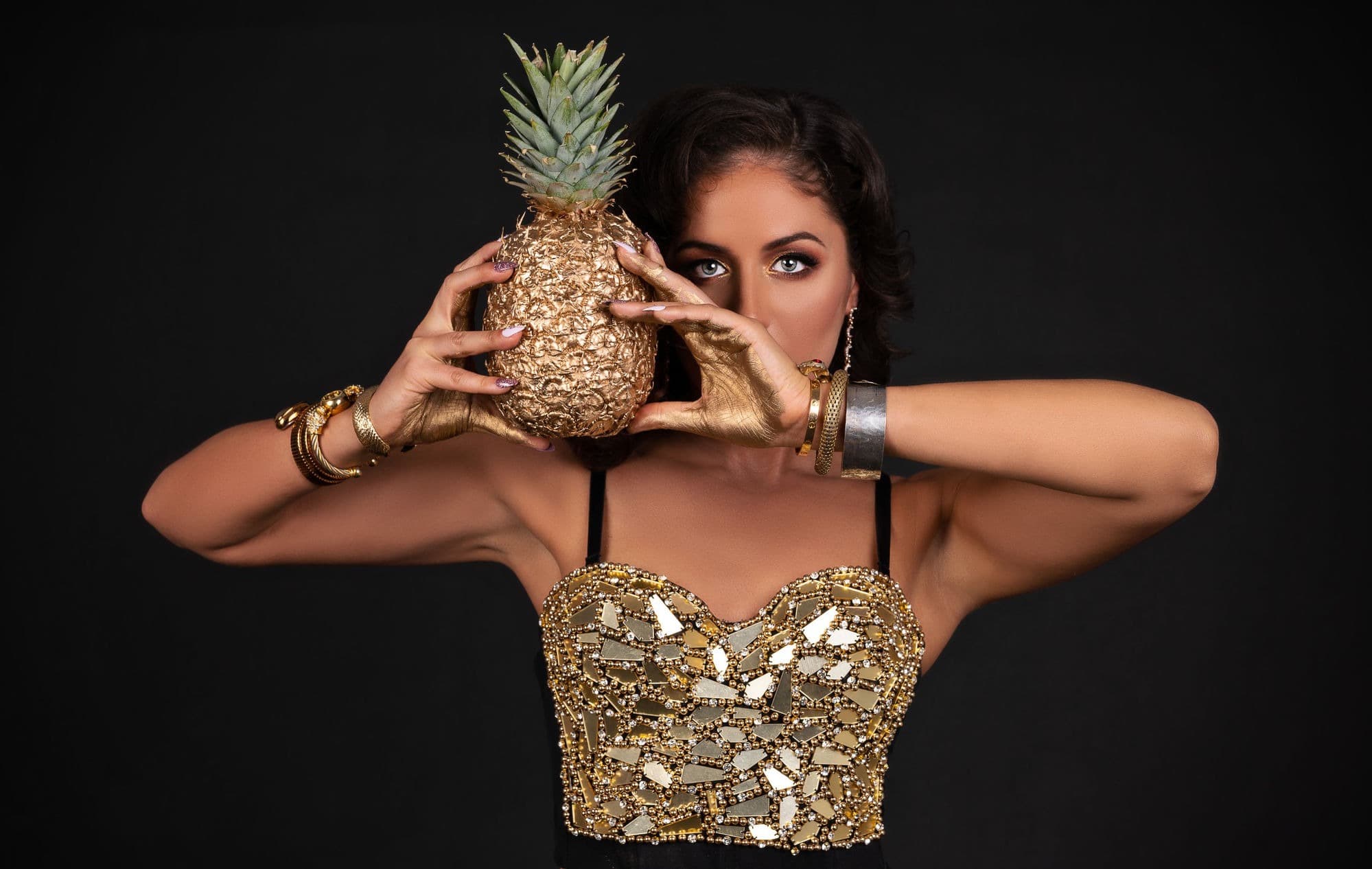 An editorial beauty shot — a woman in a gold bustier holding a gilded pineapple against black